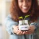 Young african american woman presenting her glass savings jar with a budding plant growing out from it at home. Happy mixed race person smiling while planning, saving and investing for her future